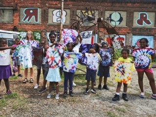 A group of children and adults holding tie-dye shirts, standing in front of an old building covered in grafiti.