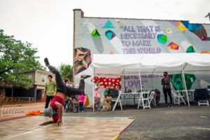 A person is break dancing in a parking lot. Behind the dancer is a group of people seated under a tent, and a colorful mural painted on the side of historic, brick building.