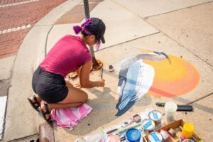 An artist kneels on the ground painting a heron and sunset on the sidewalk.