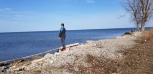 Person standing on the gravel, concrete-covered shore of the Saginaw Bay.