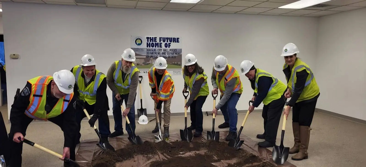 Eight Douglas city workers wearing hard hats and hi-vis vests, carrying shovels stand on a tarp covered in dirt in an office building.