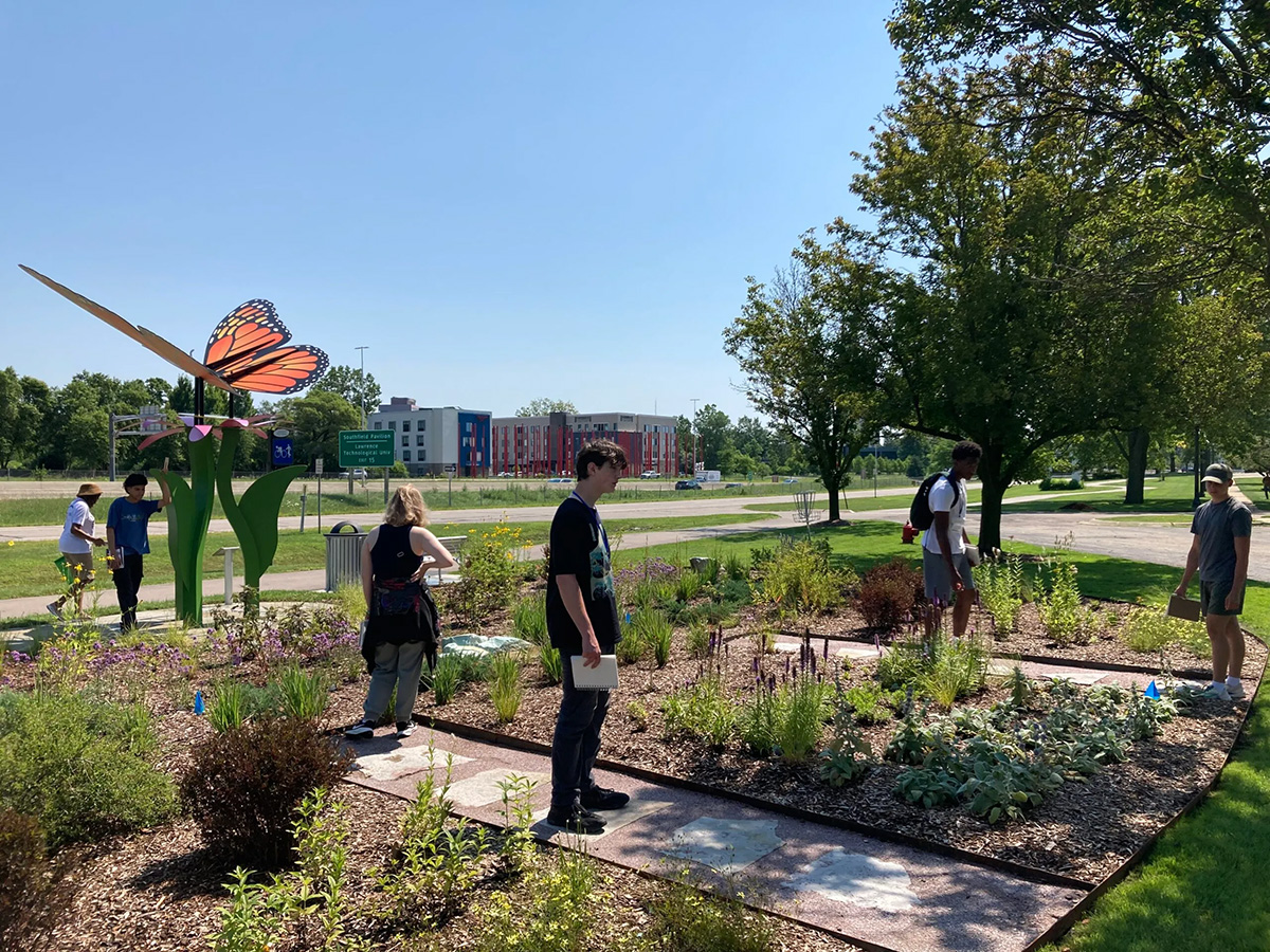 Students standing in a flower garden, there's a large Monarch Butterfly sculpture in the background.