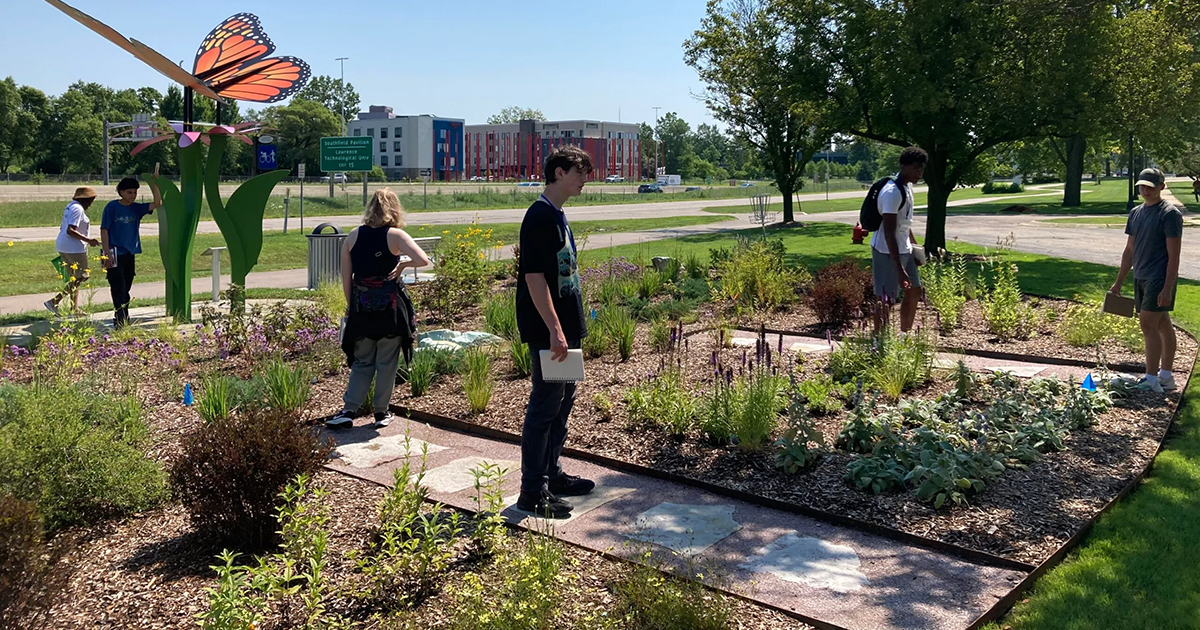 Students standing in a flower garden, there's a large Monarch Butterfly sculpture in the background.
