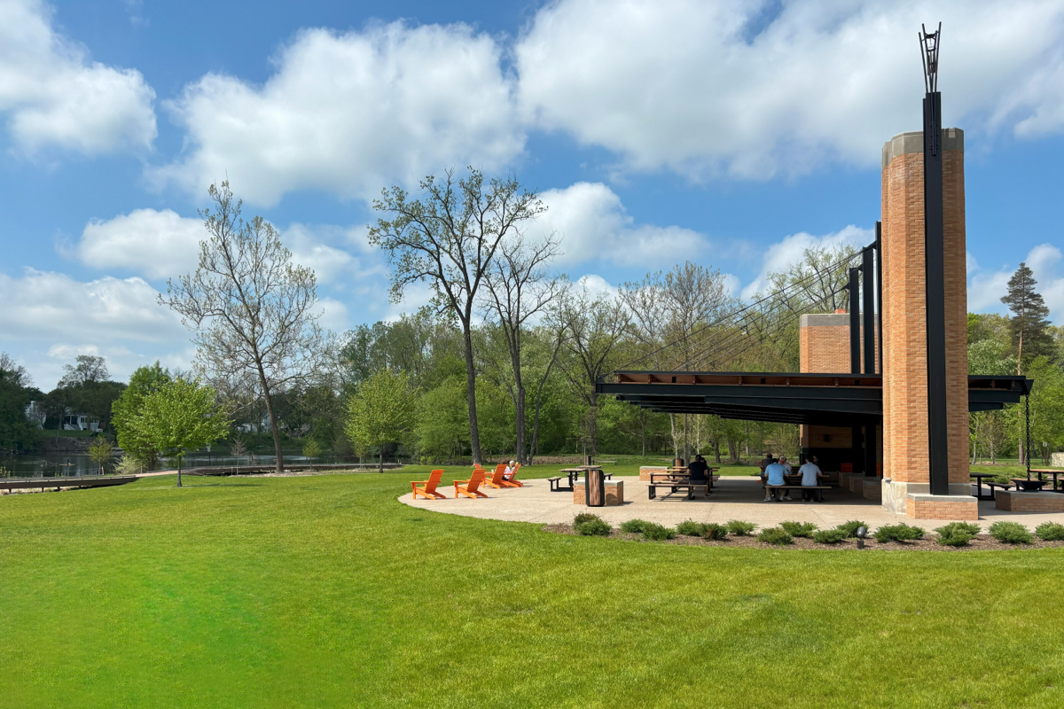People sitting under a large pavilion in the middle of a large grass field.