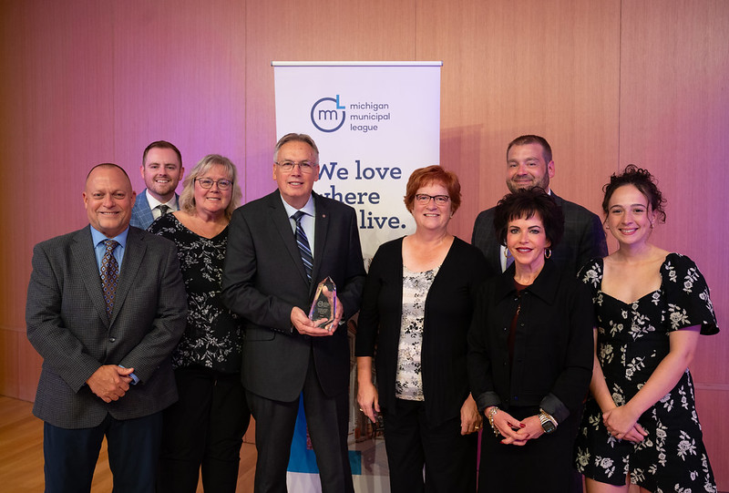 Roberty Clark holding the Michael A. Guido Leadership and Public Service Award surrounded by seven other people, standing in front of a League banner.