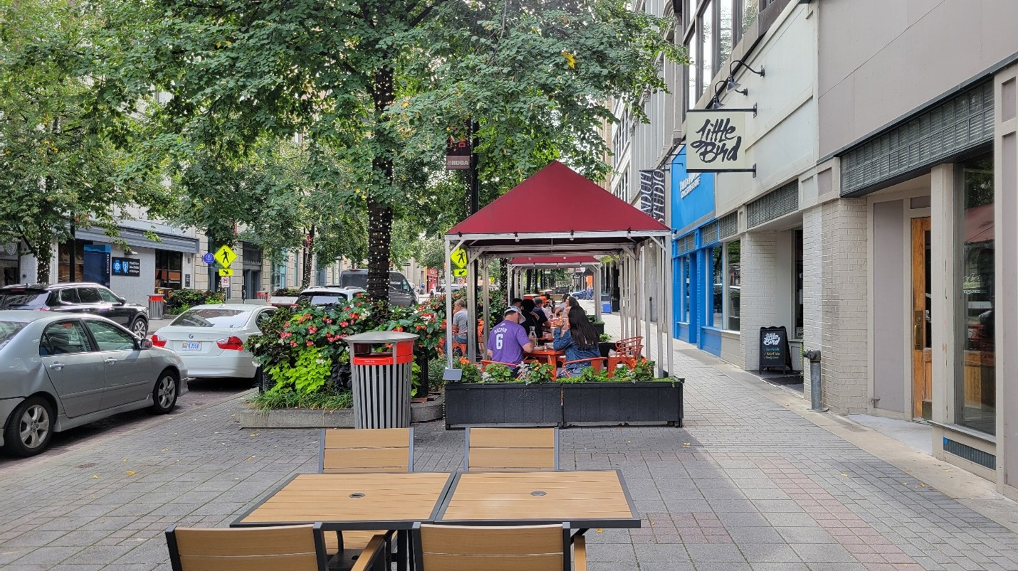 Extra wide sidewalk in Downtown Grand Rapids. There are trees, flowers, and people seated outside a restaurant.