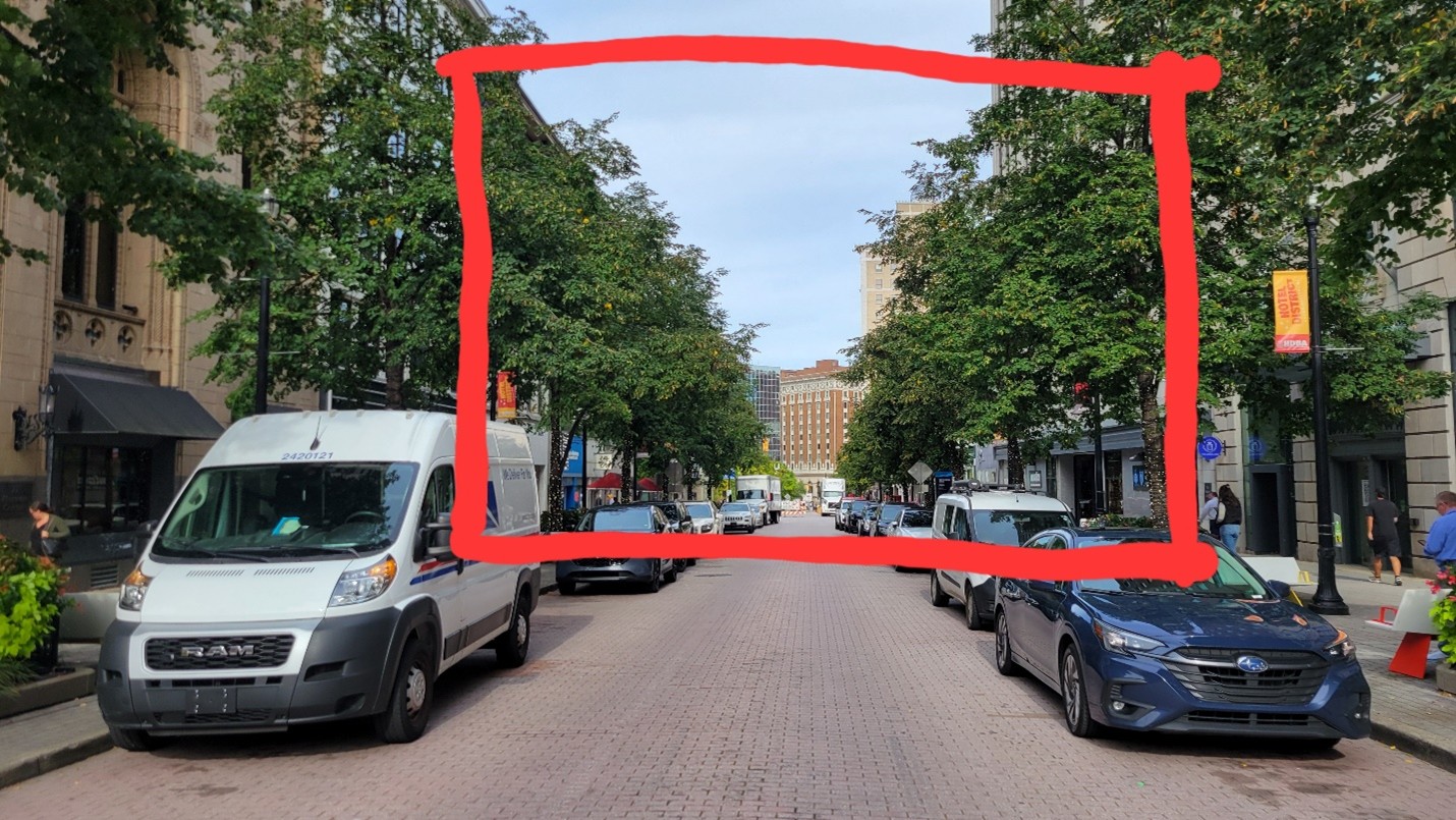 An picture taken down the center of a street in Downtown Grand Rapids. There are cars and trees lining either side of the street. There is a square drawn on the image to frame the narrow view to the end of the street.
