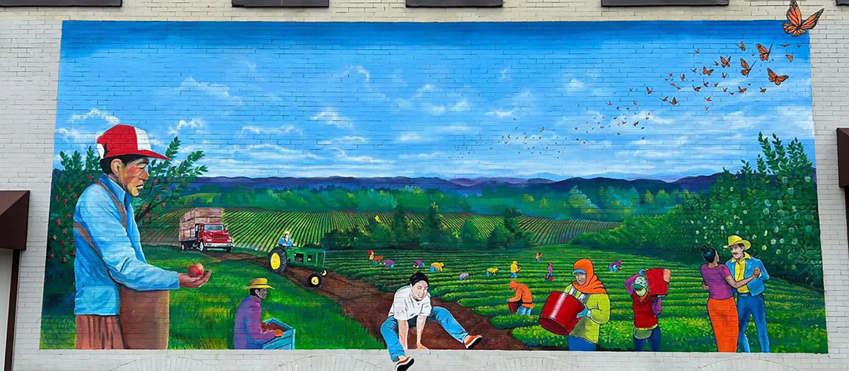 Mural on the side of a brick building, it shows hispanic people on an orchard, dancing, playing, and working.