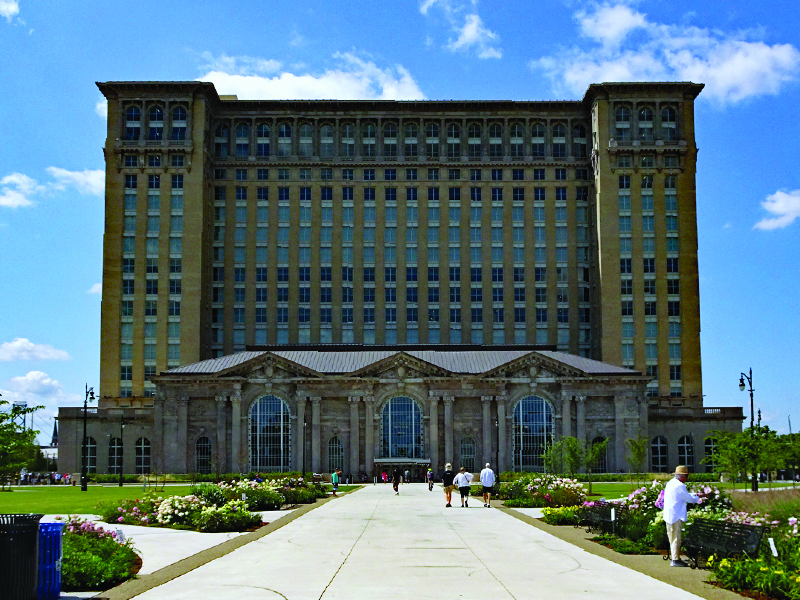 People milling about, looking at flowers in front Towering historic brick building.