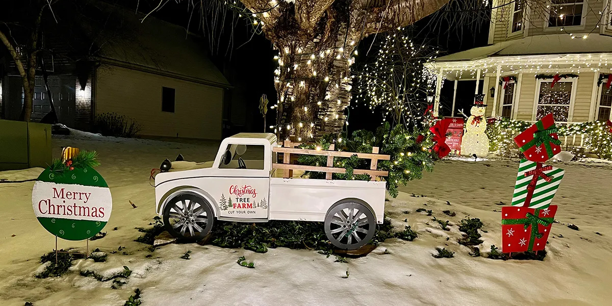 A home decorated for christmas. Along with light there is also a large model of an antique truck with the words,"Christmas Tree Farm" painted. The truck is flanked by signs, one is a bulb that says "Merry Christmas" the other looks like a stack of wrapped presents.