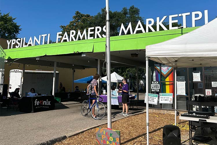 People standing in an outdoor gathering space, a large sign overhead reads, "Ypsilanti Farmer's Market."