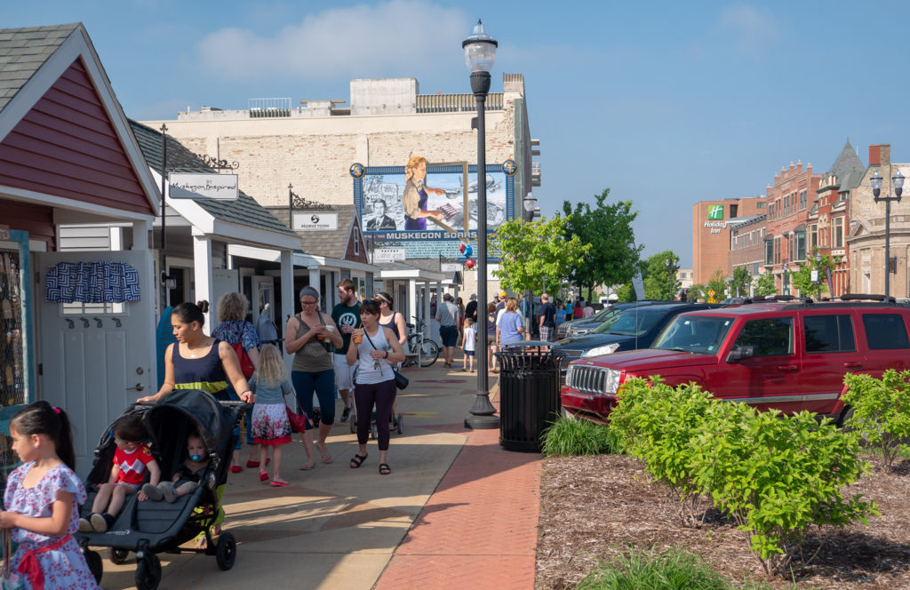 A crowd of people on a sidewalk in front of small shops in a downtown.